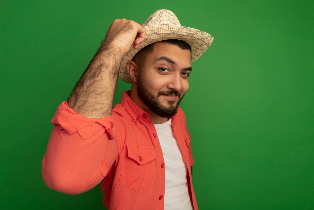 young bearded man orange shirt summer hat looking smiling confident standing green wall